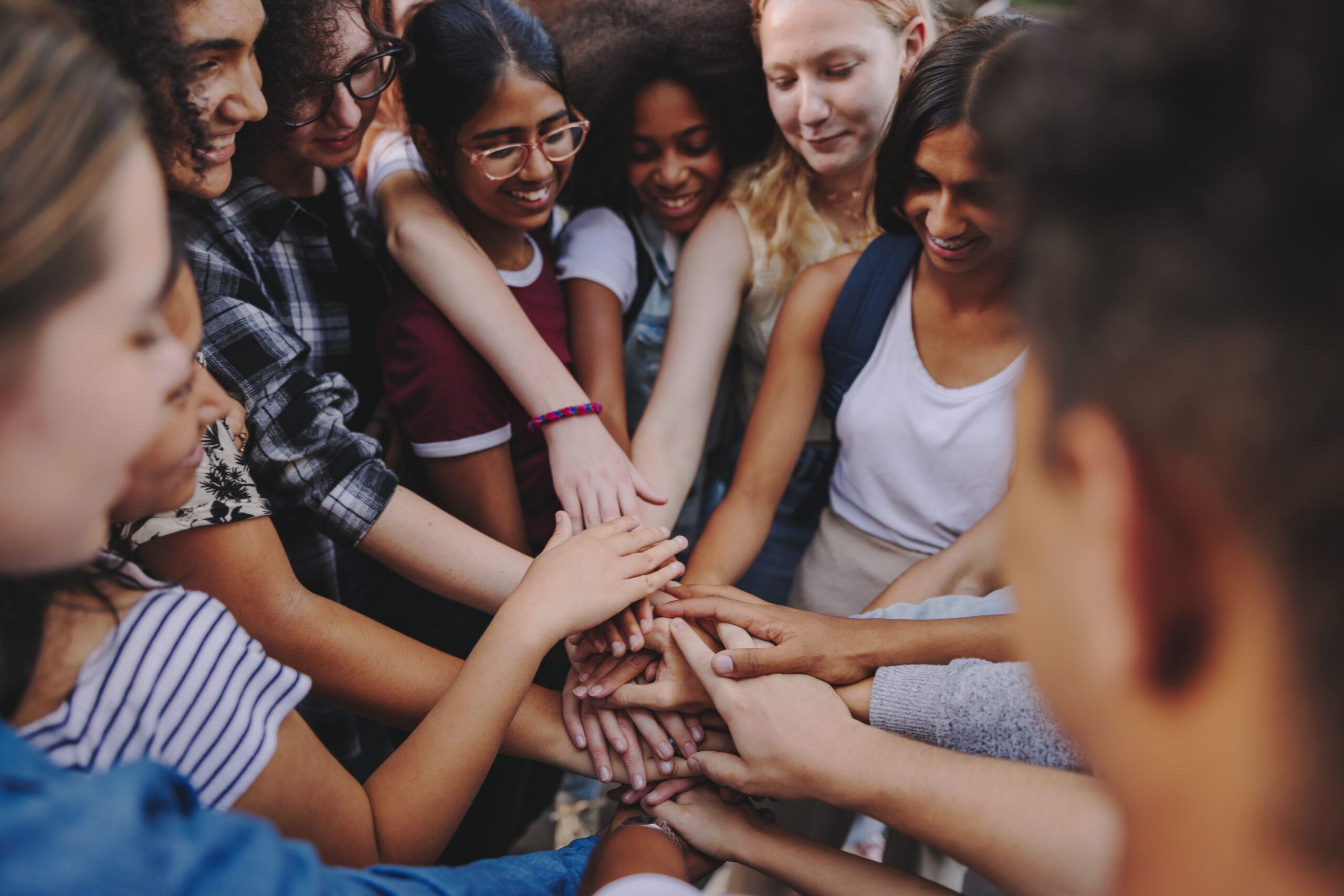 Diverse teenagers smiling happily while putting their hands together in a huddle. Group of multicultural young kids symbolizing unity and teamwork.